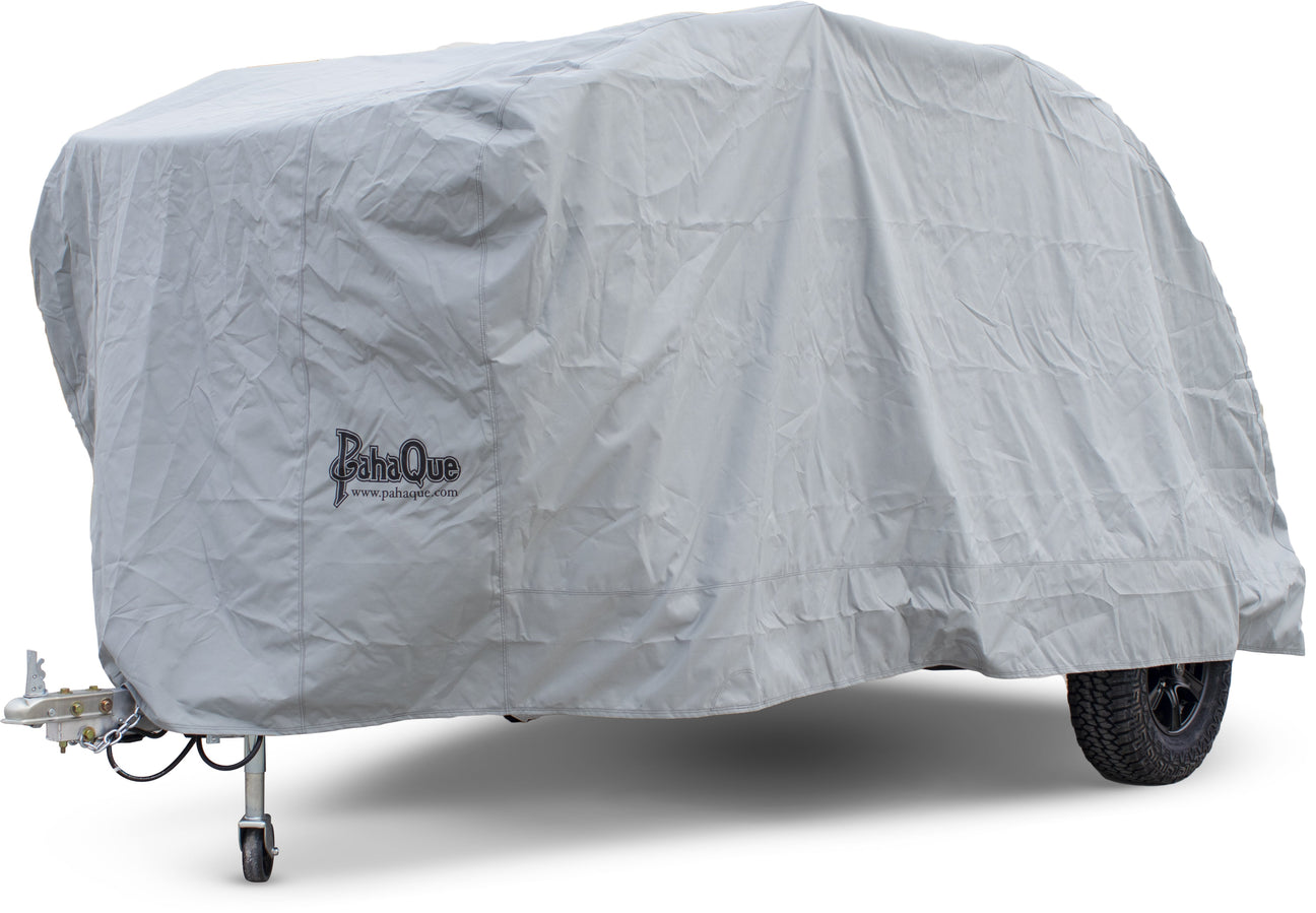 A gray protective cover is draped over a small trailer, concealing its shape and features. The cover displays the BahaQue logo and has a wheel and trailer hitch visible at the front.