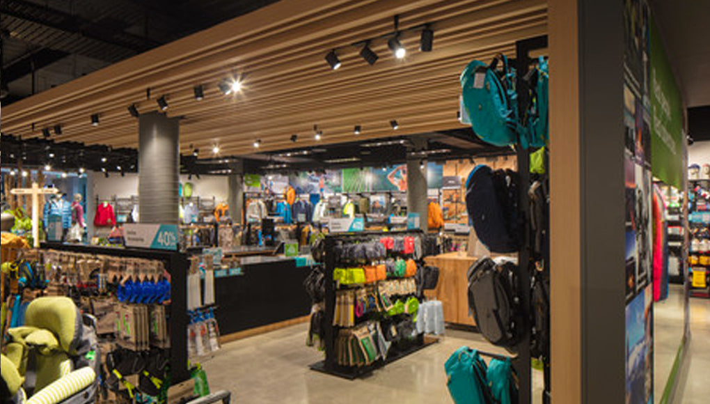 A well-lit outdoor gear store interior displays backpacks, jackets, and camping equipment on racks. Bright overhead lighting and wood accents create a modern, organized shopping environment.