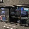A small window fitted with Paha Que Custom Black Out Curtains reveals organized shelves stocked with camping gear, sleeping bags, and containers inside a compact, gray-carpeted space for privacy.