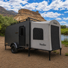 A Custom Toy Hauler Patio Ramp Tent by PahaQue Wilderness is set up by a river, attached to a black and gray camper trailer, surrounded by greenery and rocky hills beneath a partly cloudy sky.