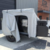 A small black camper trailer is parked outside, partially covered by the PahaQue Vega XT/AT Cover w/ Zippered Door Opening from Paha Que Custom. The light gray cover is pulled back, protecting the trailer while allowing easy access to the window and door.
