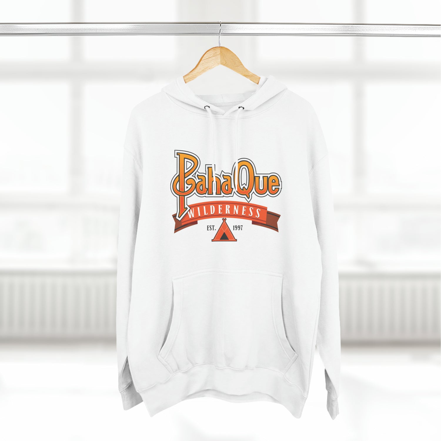 The PahaQue Unisex Premium Pullover Hoodie in white by Printify hangs on a hanger, featuring a wilderness logo with a tent graphic and Est. 1997 in red and orange tones. Its cozy fleece lining is ideal for chilly days.