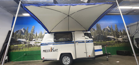 A compact white Meerkat Trailer features a blue stripe and an extended Meerkat Trailer Awning by PahaQue. Indoors, a mural of mountains, trees, and a lake adds an inviting outdoor atmosphere to the scene. Brand: Paha Que Custom.