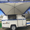 A compact white Meerkat Trailer features a blue stripe and an extended Meerkat Trailer Awning by PahaQue. Indoors, a mural of mountains, trees, and a lake adds an inviting outdoor atmosphere to the scene. Brand: Paha Que Custom.