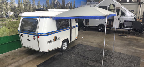 A small white Meerkat travel trailer with blue stripes is parked indoors on a tarp, with a Paha Que Custom Meerkat Trailer Awning by PahaQue set up beside it. A larger white camper sits in the background near a nature-themed mural.