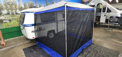 The blue-trimmed screened tent is attached to a white Meerkat Trailer indoors, set before a forest mural and other campers, highlighting the versatility of the Meerkat Trailer Awning by PahaQue from Paha Que Custom.