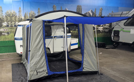 A Paha Que Custom Meerkat Trailer Side Tent for Enhanced Outdoor Adventures and Camping Comfort is set up beside a compact white trailer indoors, with another trailer and a nature mural visible in the background.