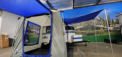 A compact Meerkat Trailer with a blue and gray Paha Que Custom Meerkat Trailer Side Tent for Enhanced Outdoor Adventures and Camping Comfort is set up indoors, backed by a mural of trees, mountains, and rocks, with exposed beams above.