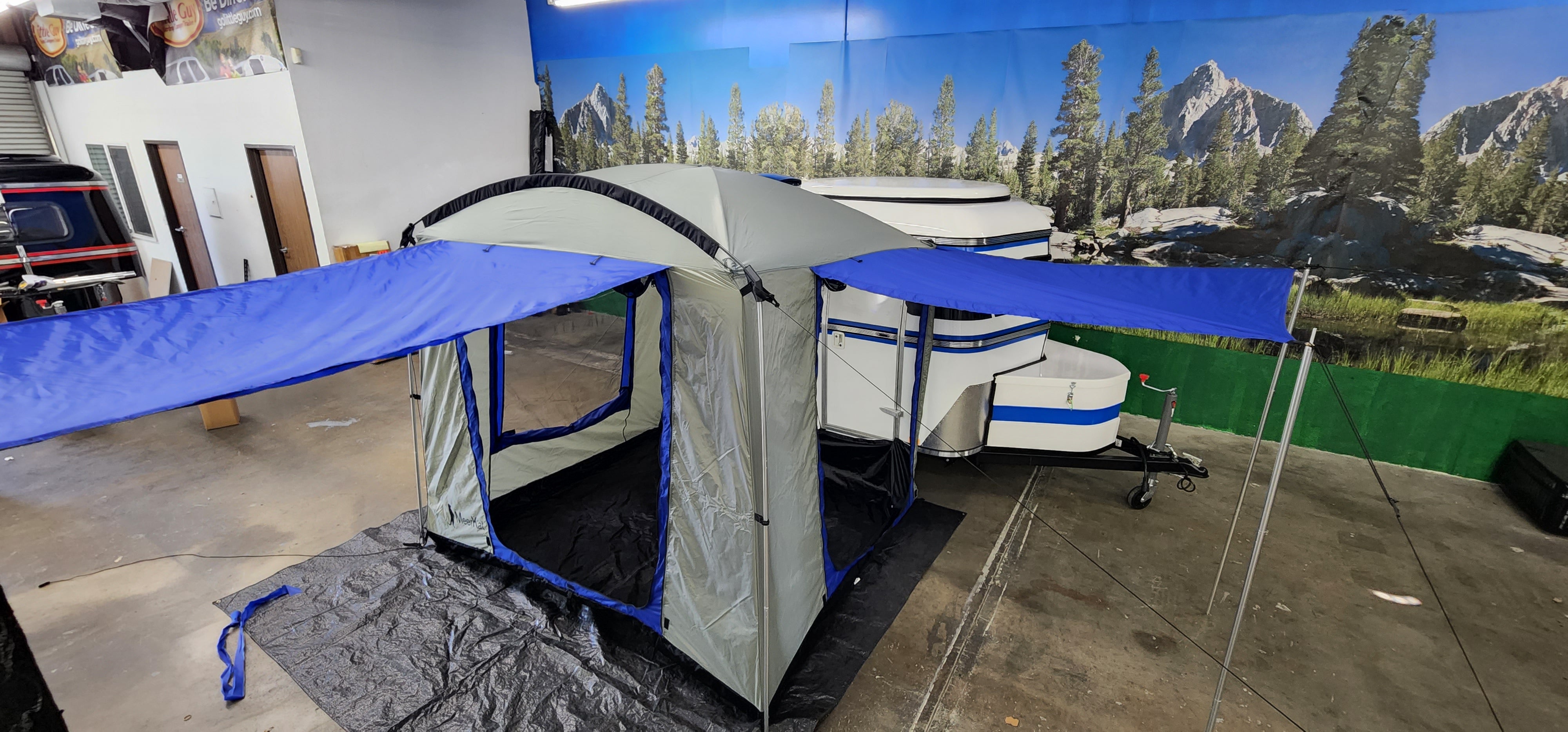 A Paha Que Custom Meerkat Trailer Side Tent for Enhanced Outdoor Adventures and Camping Comfort is set up on a black tarp indoors, next to a white and blue Meerkat trailer with a scenic forest and mountain mural in the background.