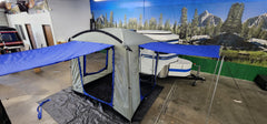 A Paha Que Custom Meerkat Trailer Side Tent for Enhanced Outdoor Adventures and Camping Comfort is set up on a black tarp indoors, next to a white and blue Meerkat trailer with a scenic forest and mountain mural in the background.
