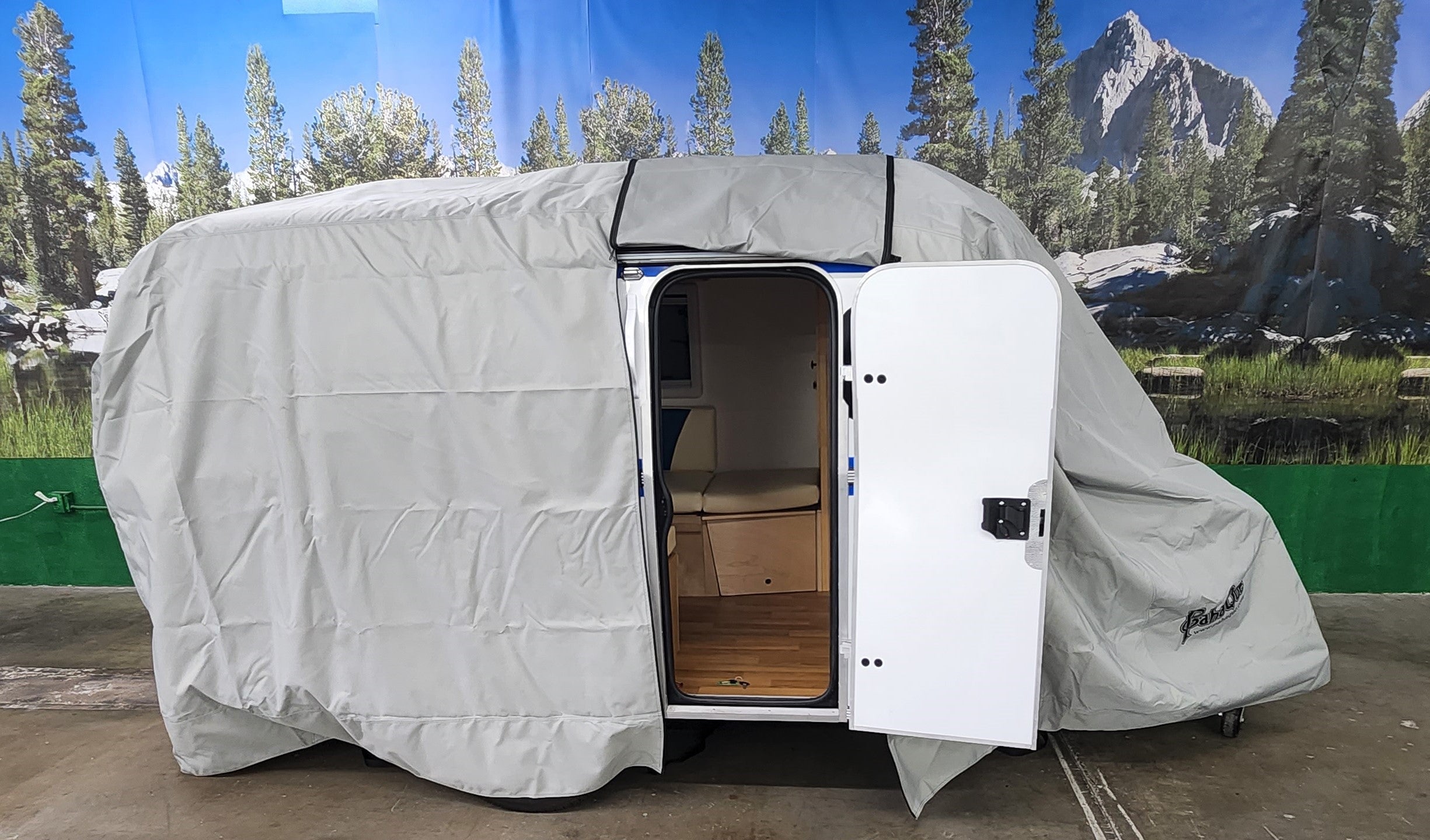 A small teardrop camper sits indoors with its white door open, showing wood flooring inside. A Paha Que Custom Meerkat Trailer Cover Heavy Duty MagnaTuff Fabric is partially removed. A forest and mountain mural is in the background.