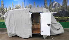 A small teardrop camper sits indoors with its white door open, showing wood flooring inside. A Paha Que Custom Meerkat Trailer Cover Heavy Duty MagnaTuff Fabric is partially removed. A forest and mountain mural is in the background.