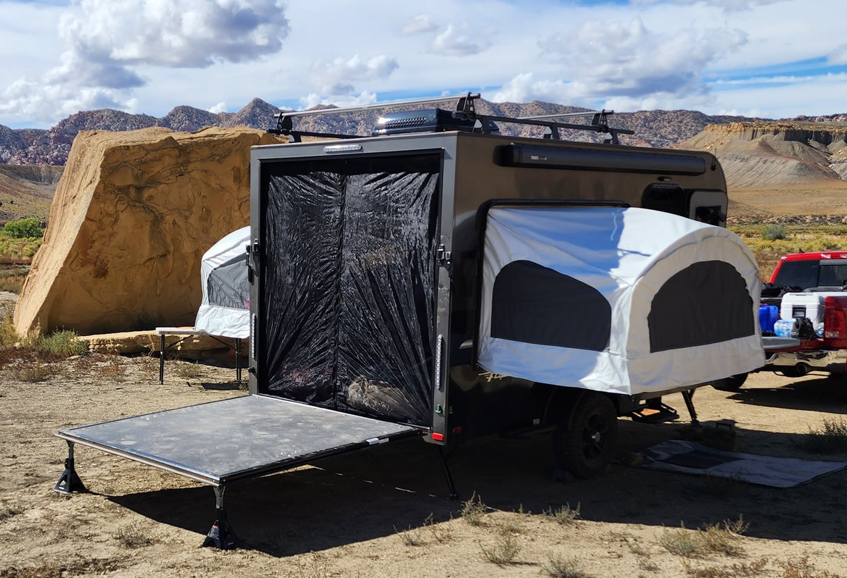 A Paha Que Custom Mesh Door/Solid Room Divider is set up on a black teardrop camper trailer with open side tents featuring No-See-Um mesh for insect protection at a desert campsite near boulders and mountains under a partly cloudy sky.