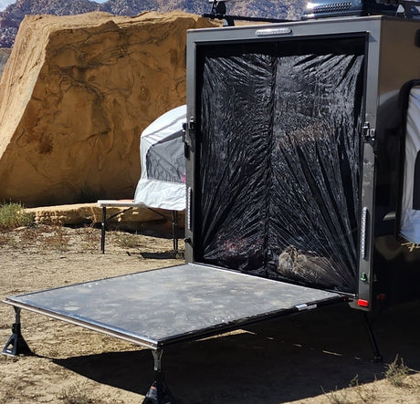 A small trailer with its back door open forms a ramp onto sandy ground in a rocky desert, protected by the Paha Que Custom Mesh Door/Solid Room Divider. A tent is visible in the background.