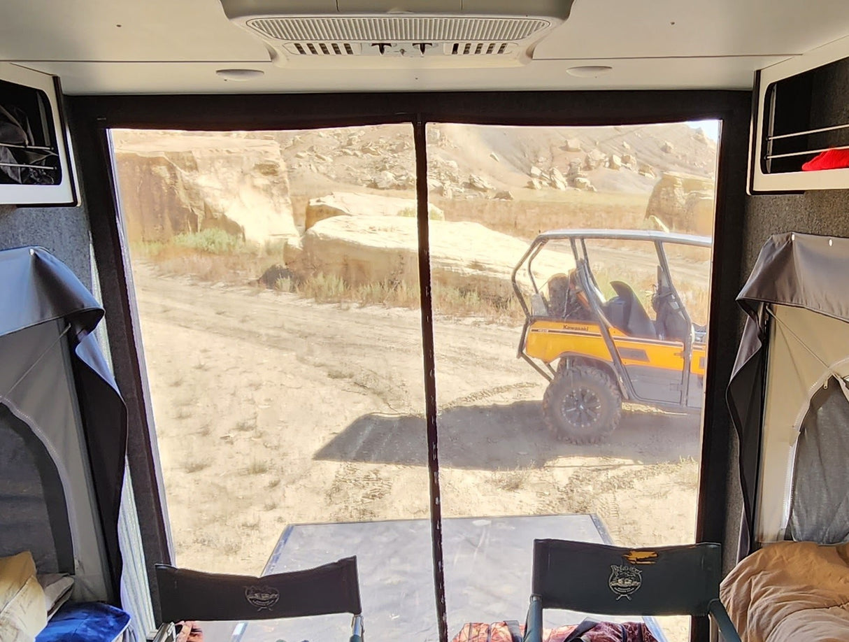 View from a camper with two sleeping areas and a table, looking out through the Paha Que Custom Mesh Door/Solid Room Divider, which offers insect protection, at a dirt road and yellow ATV among rocky terrain.