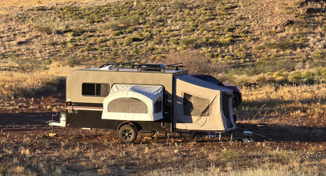 A Toy Hauler Patio Ramp Tent by Paha Que Custom for Intech Trailers is set up on dry grass in a sunny, arid landscape, with a red vehicle partially visible at the front.
