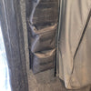 The PahaQue Wilderness Triple Gear Pouch Heavy Duty Gear Organizer is mounted on a gray carpeted wall near a curtain and two electrical switches. Mesh pockets are visible, and the floor has a black hexagonal pattern.