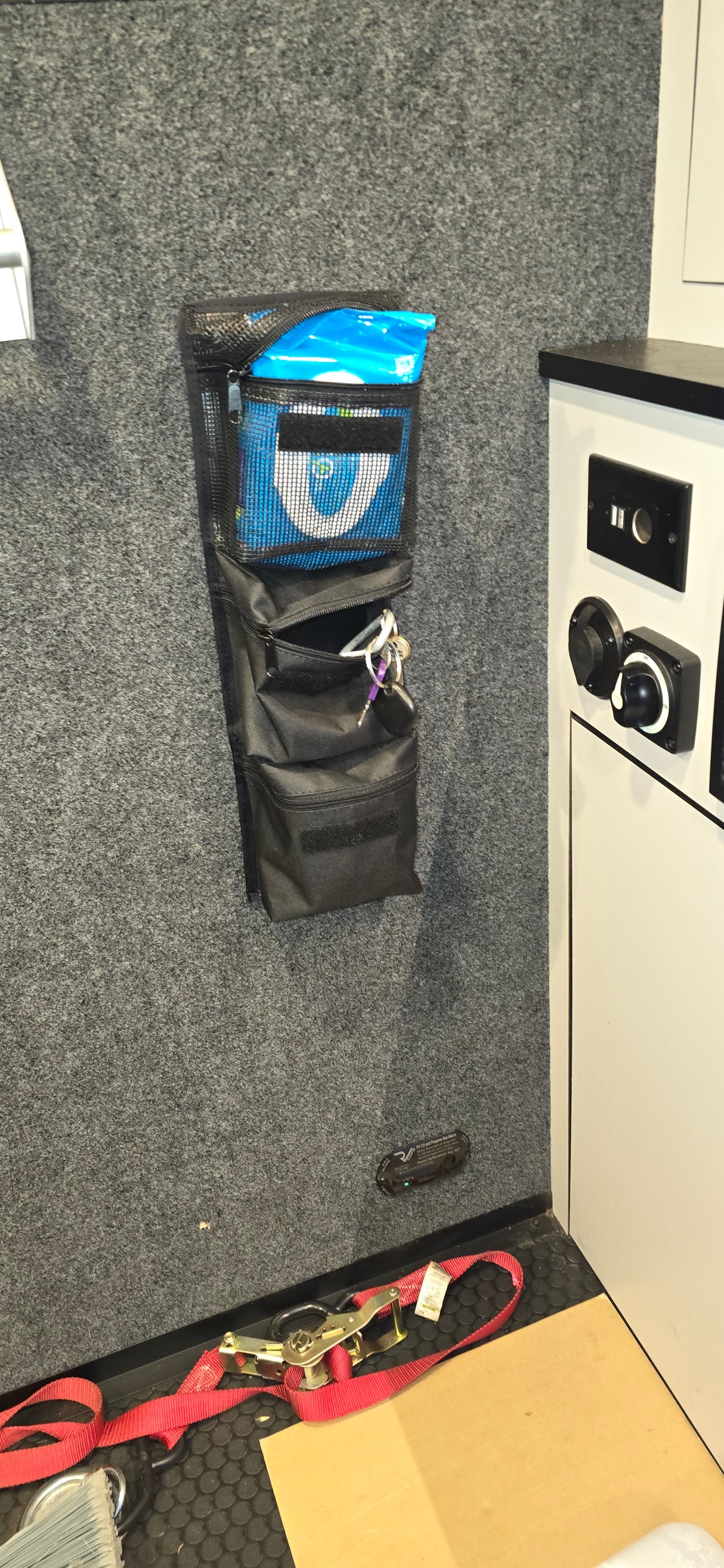 The PahaQue Wilderness Triple Gear Pouch Heavy Duty Gear Organizer for Trailers hangs on a gray wall, holding blue wipes and a key ring. Below is a folded black pouch, with switches on a white cabinet and straps scattered across the black rubber floor.