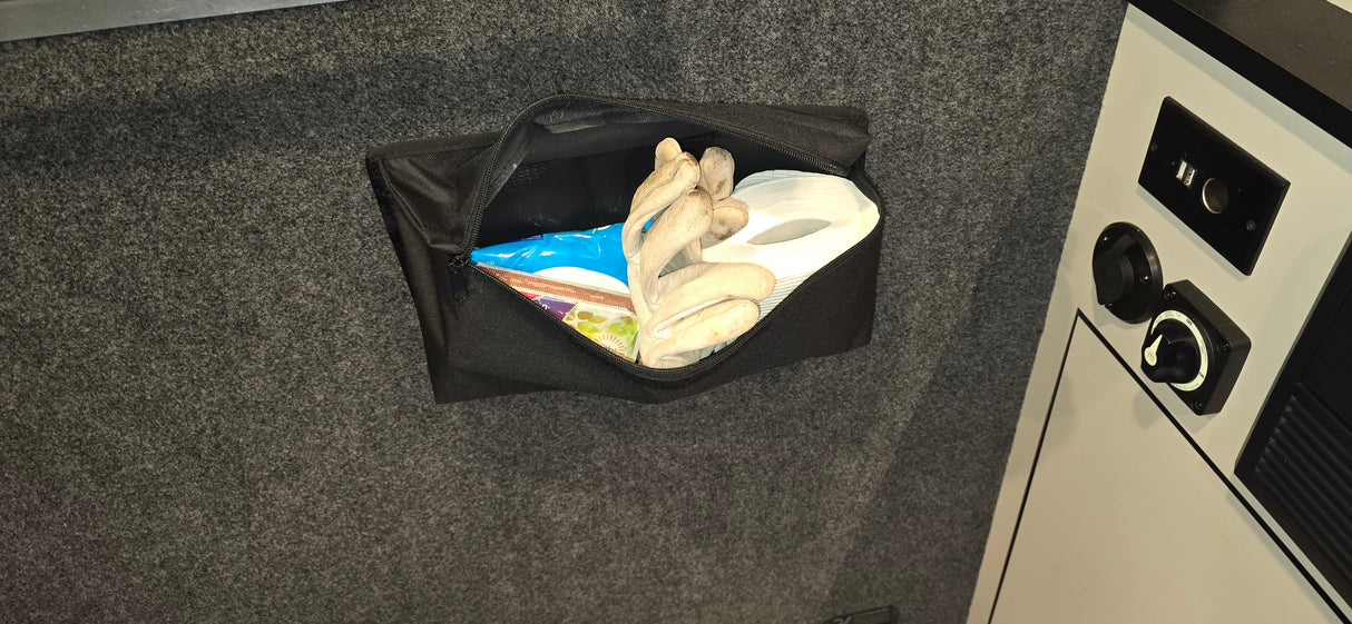 A PahaQue Wilderness PahaQue Gear Pouch is mounted on a gray wall, holding work gloves, toilet paper, and a blue package. Nearby, a white and black control panel with switches creates an efficient trailer gear storage setup.