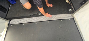 A person kneels on a black rubber floor inside an Intech trailer, pressing a fabric seam of the Toy Hauler Patio Ramp Tent by Paha Que Custom. Their face is not visible.