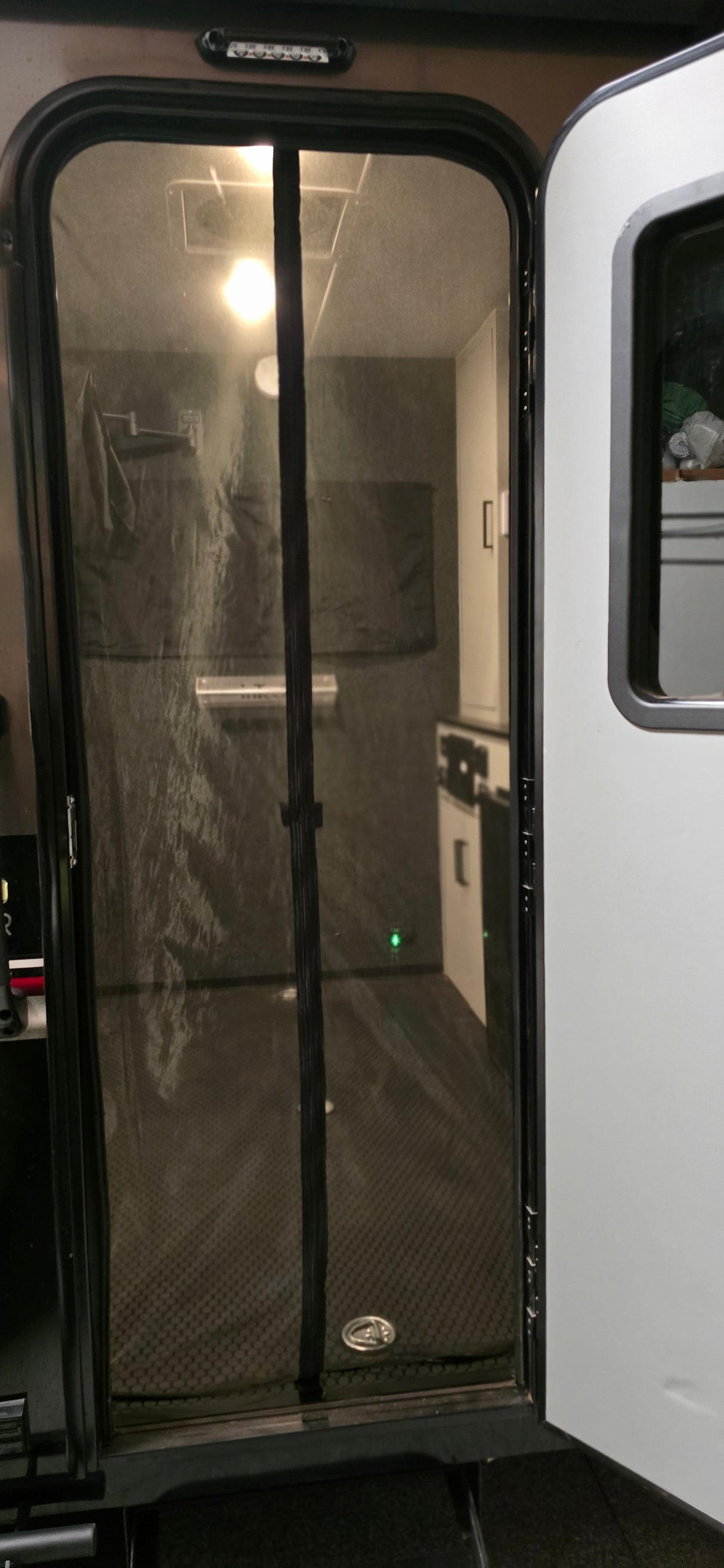Enjoy bright ceiling lights and insect protection without sacrificing your view with the Paha Que Custom Mesh Door/Solid Room Divider—peek through to a modern RV interior featuring a cabinet, counter, wall light, and towel rack.