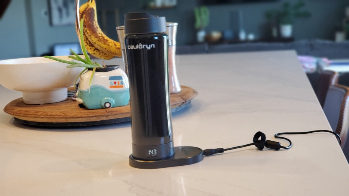 A black PahaQue Wilderness Cauldryn Variable Temperature Beverage Warmer sits charging on a kitchen counter, with bananas, a plant, and a decorative van in the background—making it an ideal sleek gift for any coffee enthusiast.