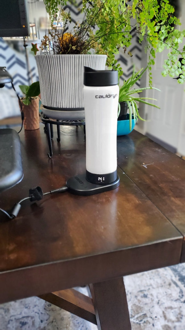 A PahaQue Wilderness Cauldryn Variable Temperature Beverage Warmer sits on a wooden desk amid green potted plants. Its base displays a number, and a computer keyboard is partially visible to the left.