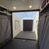 The interior of a camper or RV is shown with gray curtains on both sides and an attached, enclosed tent area at the rear, visible through the open rear door. Overhead lights are illuminated and there are storage compartments above. The setup features the Toy Hauler Patio Enclosure Ramp Tent by PahaQue Wilderness, which is custom-built to fit all Toy Hauler trailers and provides versatile options for use as either a ramp tent or an outdoor patio space, enhancing comfort and functionality.