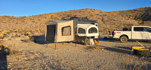 The Toy Hauler Patio Ramp Tent by Paha Que Custom for Intech Trailers is set up on rocky desert ground near a white pickup, surrounded by dry shrubs and hills beneath a clear blue sky.