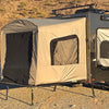 A beige Toy Hauler Patio Enclosure Ramp Tent by PahaQue Wilderness, custom built to fit all toy hauler trailers, is attached to the side of a dark camper trailer, set up on rocky desert ground with hills and sparse vegetation in the background.
