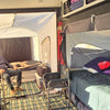 A man smiles inside a spacious camper with two elevated beds, checkered floor mat, camping chairs, and bedding as sunlight pours through an open PahaQue Wilderness Custom Toy Hauler Patio Ramp Tent for outdoor adventures.