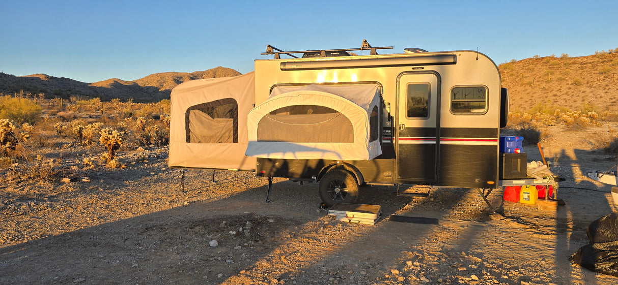 The Toy Hauler Patio Ramp Tent by Paha Que Custom for Intech Trailers is set up in a rocky desert at sunset, surrounded by sparse vegetation and hills, with outdoor gear beside it and weatherproof tent sections popped out.