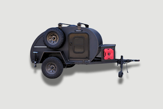 A compact black teardrop camper with off-road tires, roof rack, spare tire, and a red gas can is shown on a light gray background. Protect it with PahaQue Wilderness Teardrops Northwest Trailer Covers for all-weather durability.