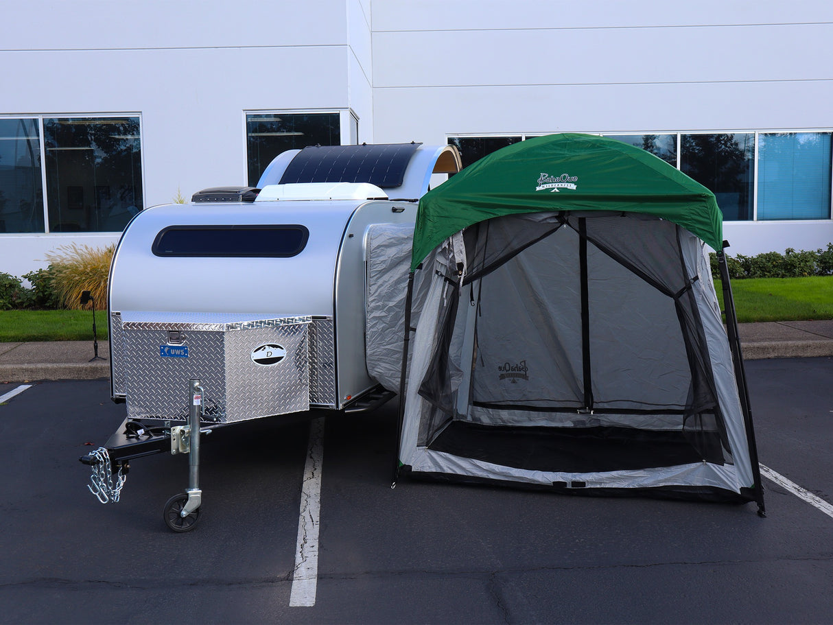 A Paha Que Custom 5x7 Trailer Side Tent Screen Room for Camping and Outdoor Adventures is set up next to a white teardrop trailer by a green-roofed canopy, with modern white buildings and landscaped greenery in the background.