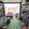 Interior of the Paha Que Custom Toy Hauler Patio Ramp Tent by PahaQue for Intech Discover Trailers, featuring three beds with blankets and personal items, two chairs, a small table, plaid flooring, and sunlight from the open rear area.