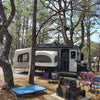 A pop-up camper is set up among pine trees at a campsite. Nearby, a PahaQue Wilderness Custom Toy Hauler Patio Ramp Tent for Outdoor Adventures, folding chairs, a fire pit, pink flamingo decor, and an outdoor mat complete the peaceful scene.