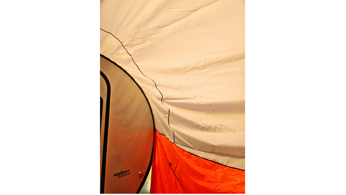 Close-up of a beige, orange, and white tent showing its fabric texture and seams, with part of a NuCamp TAB 360 Trailer Awning by Paha Que Custom and its teardrop camper visible on the left.