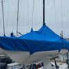 A Soling sailboat, covered by a Paha Que Custom Soling Sailboat Storage Cover, is docked on land among other boats beneath overcast skies.