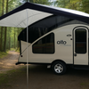 The Alto Canopy by Paha Que Custom, a modern teardrop-style camper with a waterproof canopy, is parked on a forest path surrounded by tall trees and lush greenery.