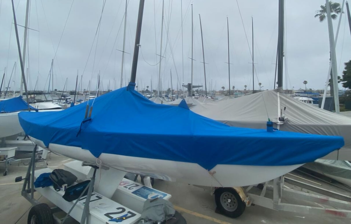 Several Soling sailboats are docked on land, their sails and cockpits protected by Paha Que Custom Soling Sailboat Storage Covers. Other boats and masts are visible in the marina under a cloudy sky.