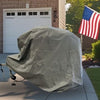 A vehicle with a gray Paha Que Custom Ushi Chuck MAX Trailer Cover by PahaQue Heavy Duty Weather Proof Protection is parked in a driveway near a garage. An American flag stands by the lawn, with trees and shrubs behind.