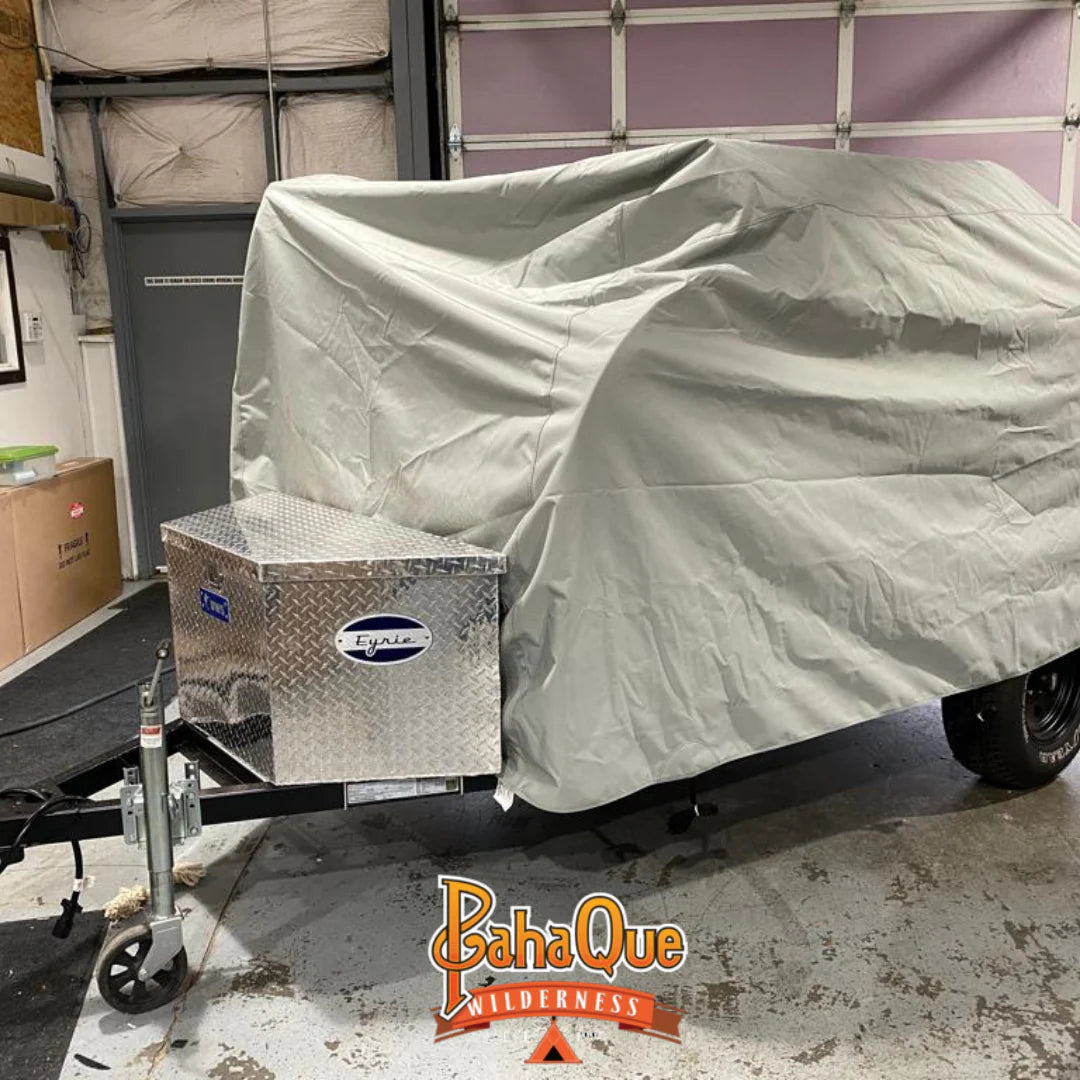 A small trailer with a durable Aero Teardrops Trailer Cover by PahaQue Wilderness made from MagnaTuff fabric is parked in a garage. A diamond-plate storage box is attached to the front, and the PahaQue Wilderness logo appears at the bottom.