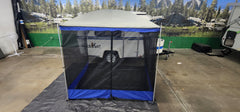 A blue and gray screened tent is pitched on a tarp indoors near a small white Meerkat Trailer, featuring the Meerkat Trailer Awning by PahaQue from Paha Que Custom. A forest and mountain mural forms the scenic background.