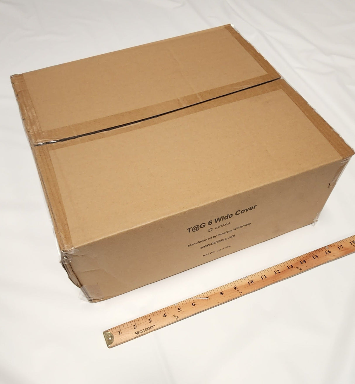 A closed cardboard box labeled OUT108 Hiker 5x10 Trailer Cover by Paha Que Custom, possibly containing a waterproof trailer cover, sits on a white surface next to a wooden ruler, showing the box is about 14 inches wide.