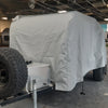 A PahaQue Wilderness Colorado Teardrops Canyonland Pinnacle Trailer Cover in gray MagnaTuff fabric shelters a small trailer with a large spare tire and metal storage box at the front, parked inside a building with a partially exposed ceiling.