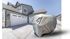 A large, light gray Paha Que Custom Meerkat Trailer Cover Heavy Duty MagnaTuff Fabric conceals a trailer in the driveway of a modern two-story house with green siding and three white garage doors under a clear sky.