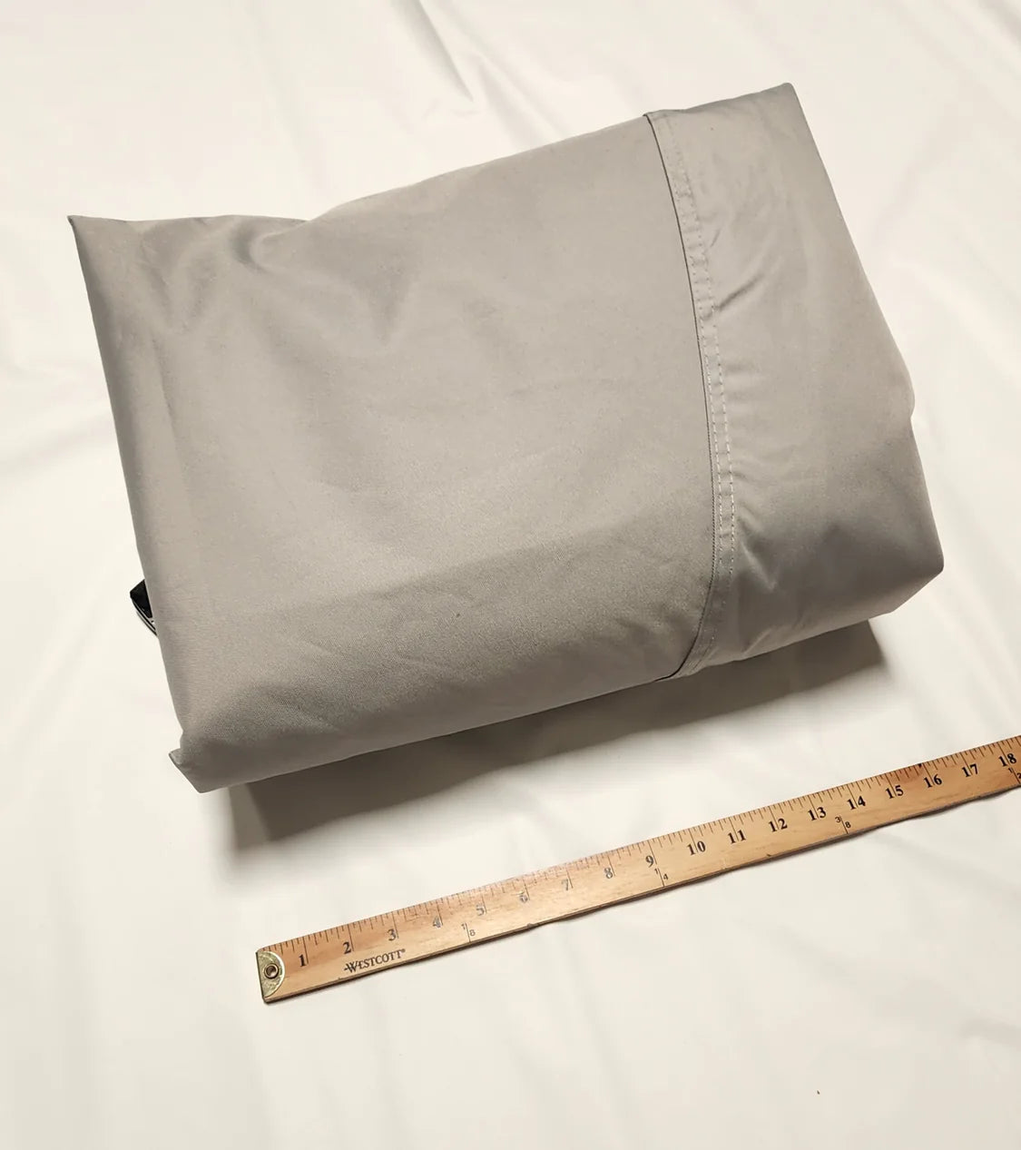 A folded NuCamp TAB 320 Outback/Boondock Trailer Cover by PahaQue, in light gray fabric from Paha Que Custom, is lying on a white surface beside a wooden ruler for scale.