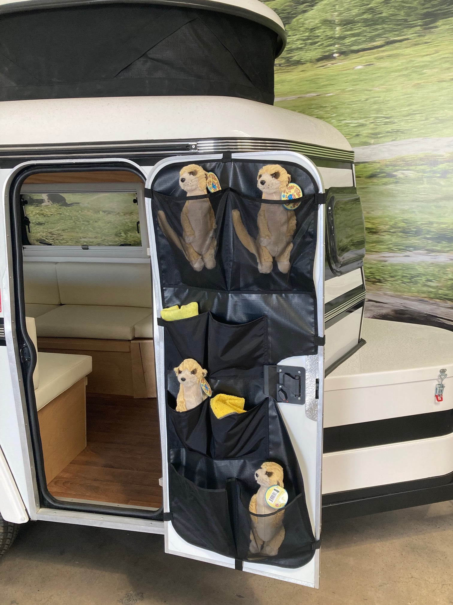 Stuffed meerkat toys sit in custom pockets of the PahaQue Wilderness Meerkat Door Kaddy, hanging on the open door of a small white camper. The light wood interior contrasts with the outdoor backdrop beyond the Meerkat Trailer storage.