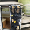 Stuffed meerkat toys sit in custom pockets of the PahaQue Wilderness Meerkat Door Kaddy, hanging on the open door of a small white camper. The light wood interior contrasts with the outdoor backdrop beyond the Meerkat Trailer storage.