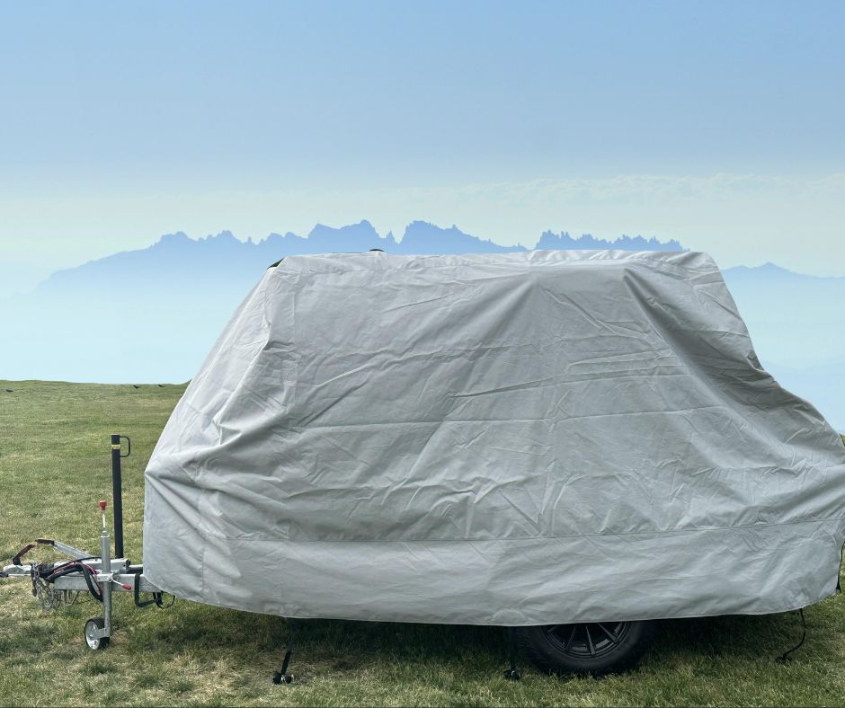 A small trailer sits on grass beneath a clear sky and hazy mountains, covered with the Hero Trailer Cover by Paha Que Custom, featuring heavy duty MagnaTuff fabric protection.